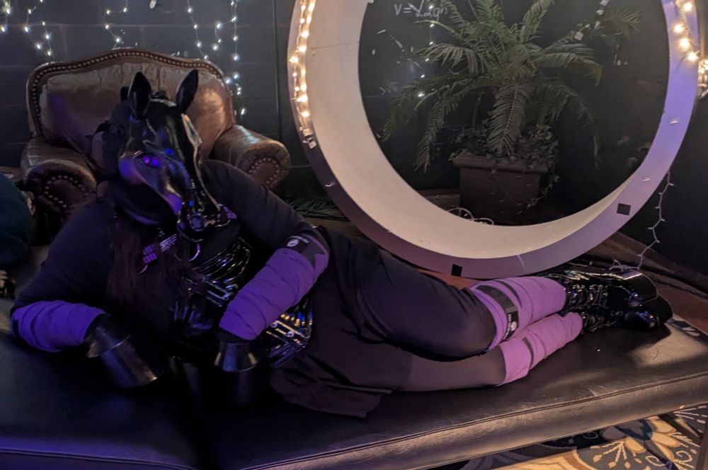 Nyxie in a black and purple pony getup, purple wraps and a PVC corset and boots with 3d printed mask and hooves, lounging on a piece of dungeon furniture. 