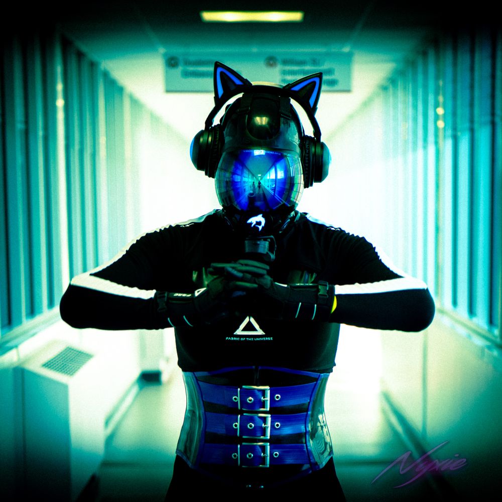 A black and blue techwear clad drone posing fairly menacingly, the green hallway background reflects nicely on their blue visor. They look ready to strike.