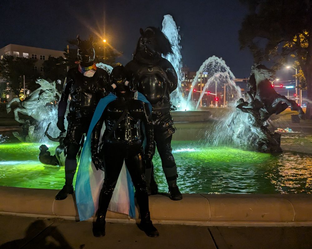Three rubber clad figures standing in front of a green lit fountain with horse statues behind it. 

From left to right its: a latex pony player with a pride rainbow bandana
Center is a latex clad femme drone , with a trans pride flag like a cape and rubber and trans pride inserts on their gas mask. 
Right: an all black pony player with a leather mask and a haired main