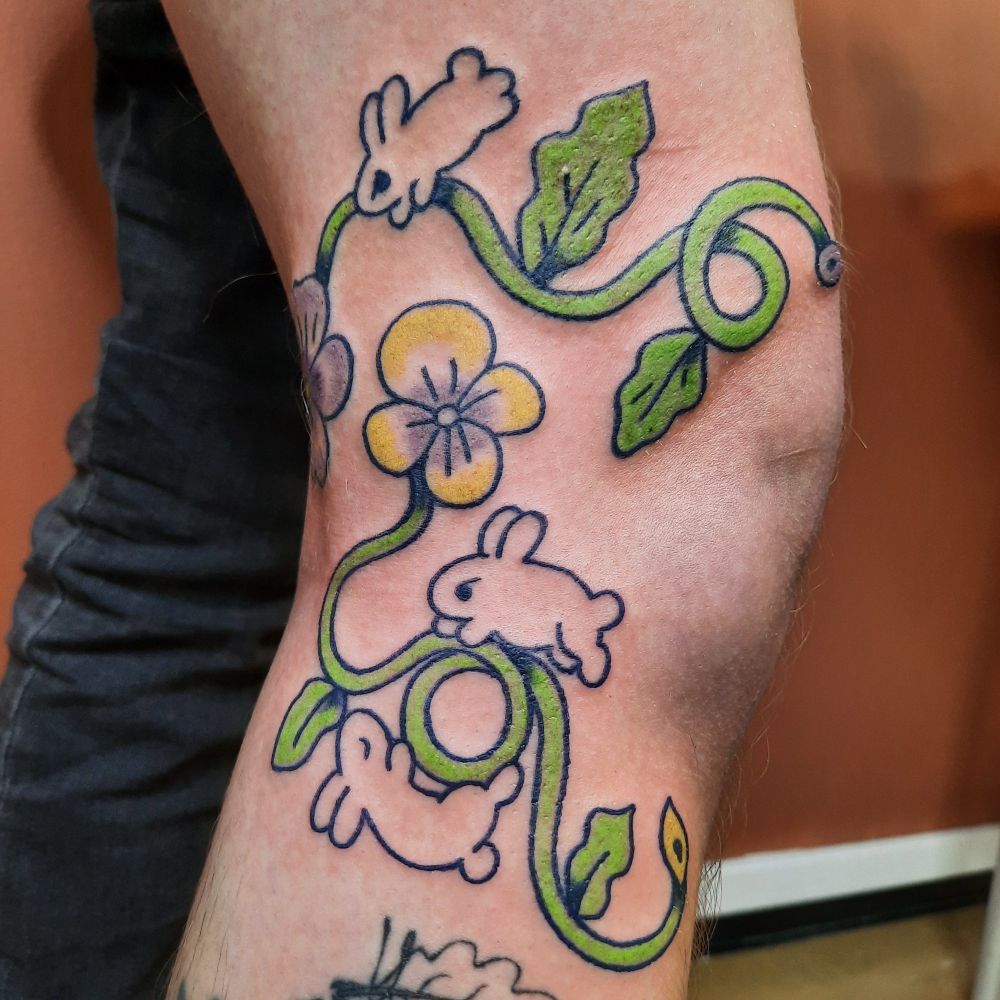 Color tattoo of two pansies with long curly stems. One flower is yellow and other purple. There are also three bunnies tattooed next to the flowers. The tattoo is done on side of the knee.