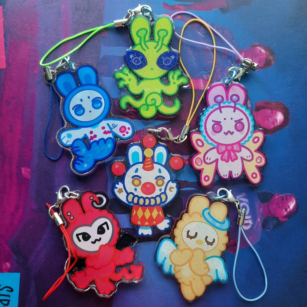 Photo of six different colorful acrylic charms with cell phone straps. All the characters resemble a bunny. One is a mermaid bunny, alien,  moth, clown, devil and angel.