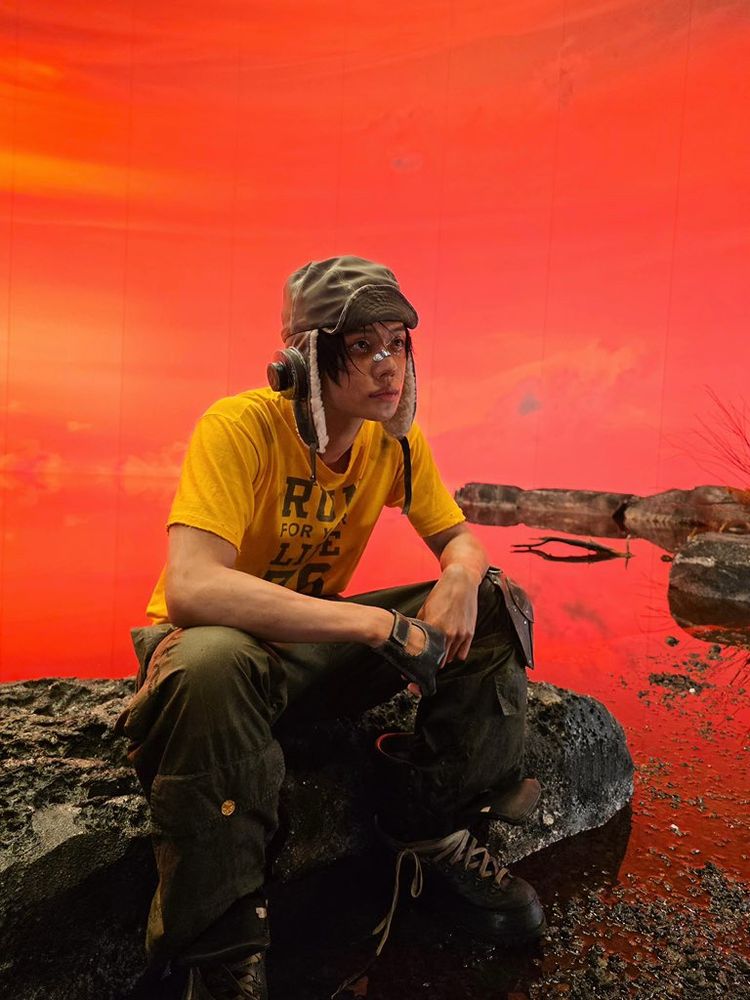 Behind-the-scenes Afterglow concept YJ. He’s just sitting on a rock in his concept outfit, facing right, & staring out into space 