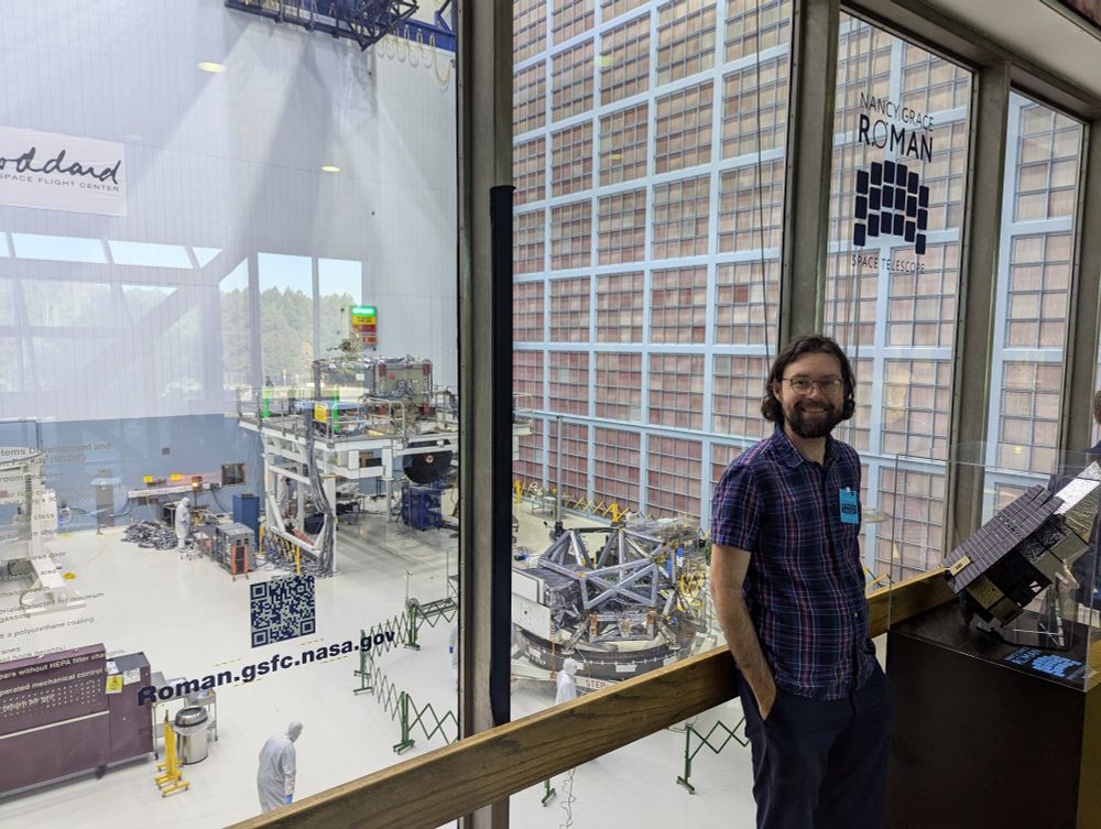 A photo of a smiling white man with dark hair and a beard leaning against a window. Behind the window is a huge cleanroom holding pieces of spaceflight hardware. A large sticker on the window reads "Nancy Grace Roman Space Telescope."