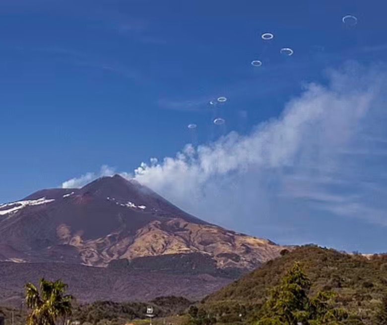 Photo of Mt. Etna showing a white cloud drifting away from the peak along with a series of rings.