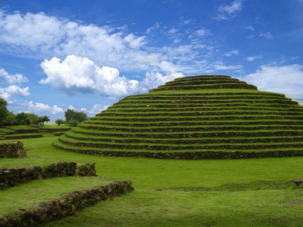 Photo of one of the circular pyramids. Rising from the ground, the pyramid has 17 green grassy/mossy steps. The top 4 steps are considerably smaller than the lower steps, resulting in a flat, grassy area. Low stone walls from one of the platforms is seen in the lower left corner of the photo. Everything in the picture, except the vertical walls and steps, are covered with bright green grass/moss.