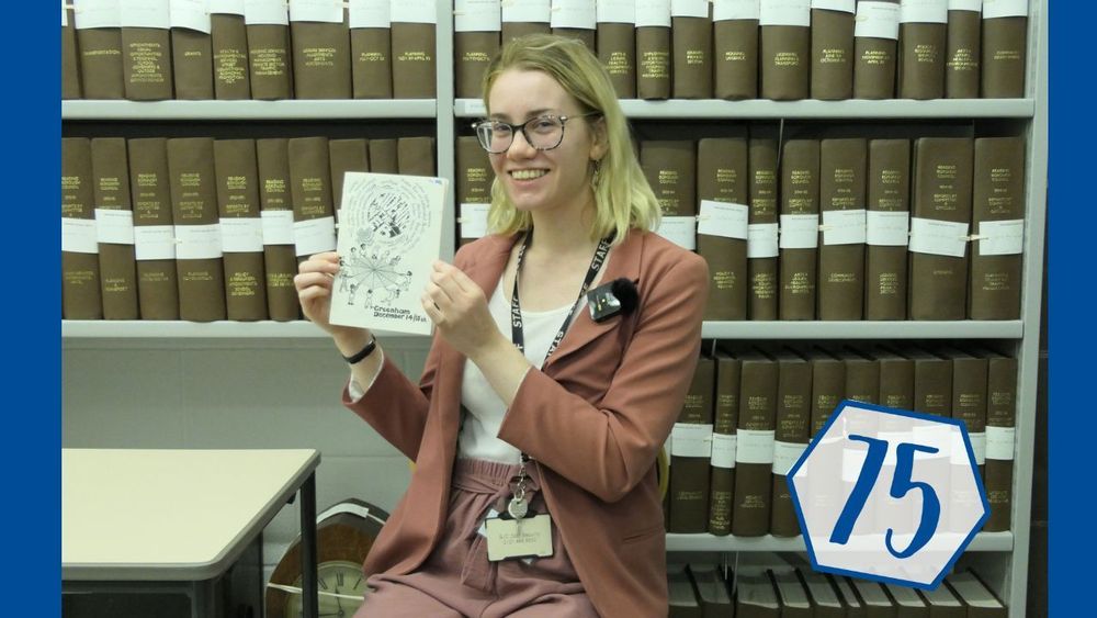 A women holding a pamphlet sitting in front of rows of books.
