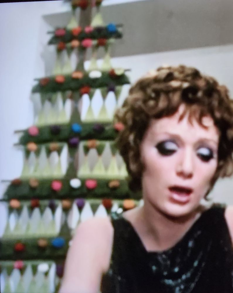 Fancy Italian lady sits in front of a floor-to-ceiling Christmas tree crafted from yarn, string & wooden sticks 