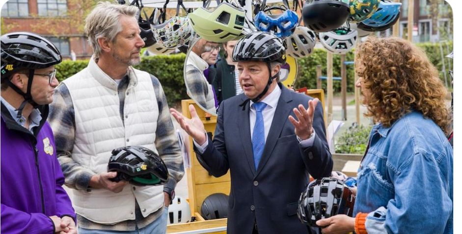 Minister Madlener staat geanimeerd met een pak aan en een fietshelm op aan drie mensen met fietshelmen op of in de hand te praten. Aan een koord in de lucht hangt een rijtje fietshelmen