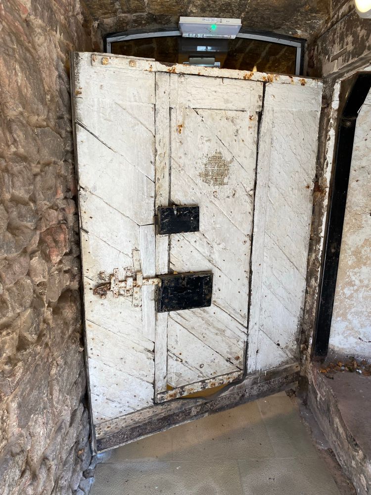Photograph of the inside face of a large, almost square-plan timber doorway, ajar in its masonry doorway. The door contains a plain wicket set centrally, with two heavy iron box locks on the wicket. The wicket and main door both have an inner skin of boards set diagonally rather than the conventional horizontal. This is rare but not unheard of. The image is of the main door to the stair tier at the Town House or Tollbooth of Dunbar in East Lothian (Scotland).