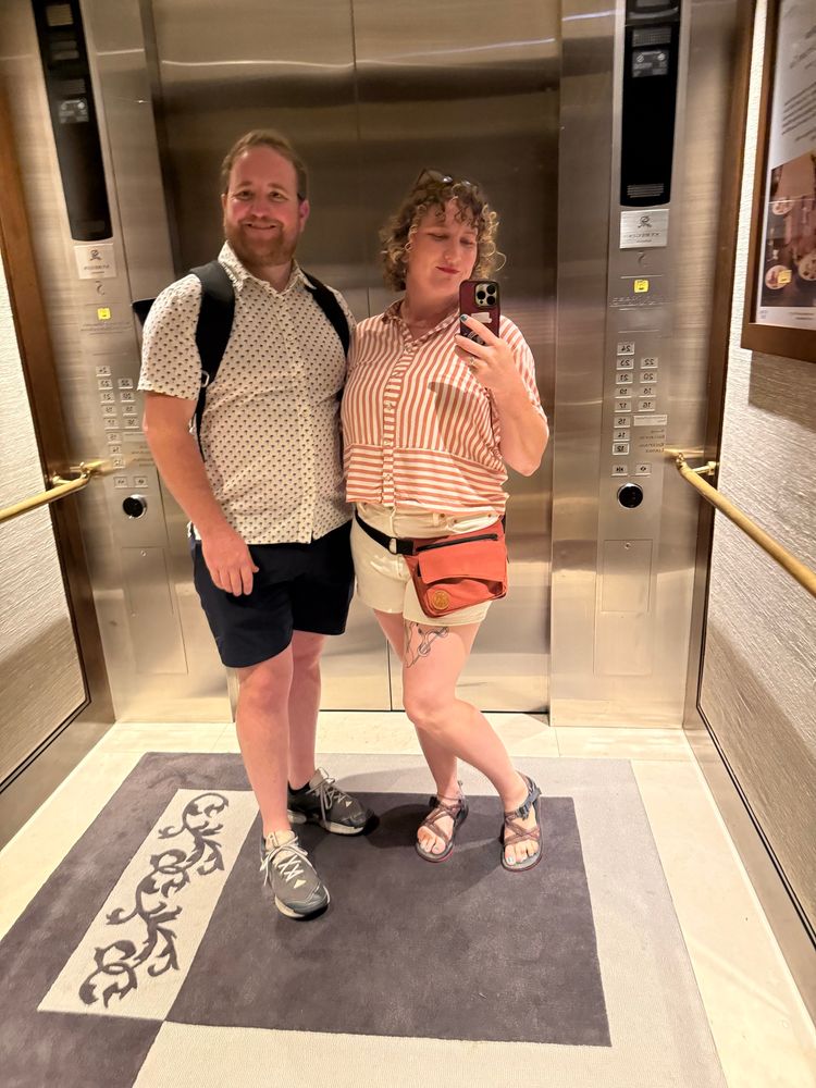 A man and woman in an elevator. The man wears a light short sleeved button up shirt, dark shorts and attic shoes. The woman wears and pink and white striped shirt and white shorts and sandals. 