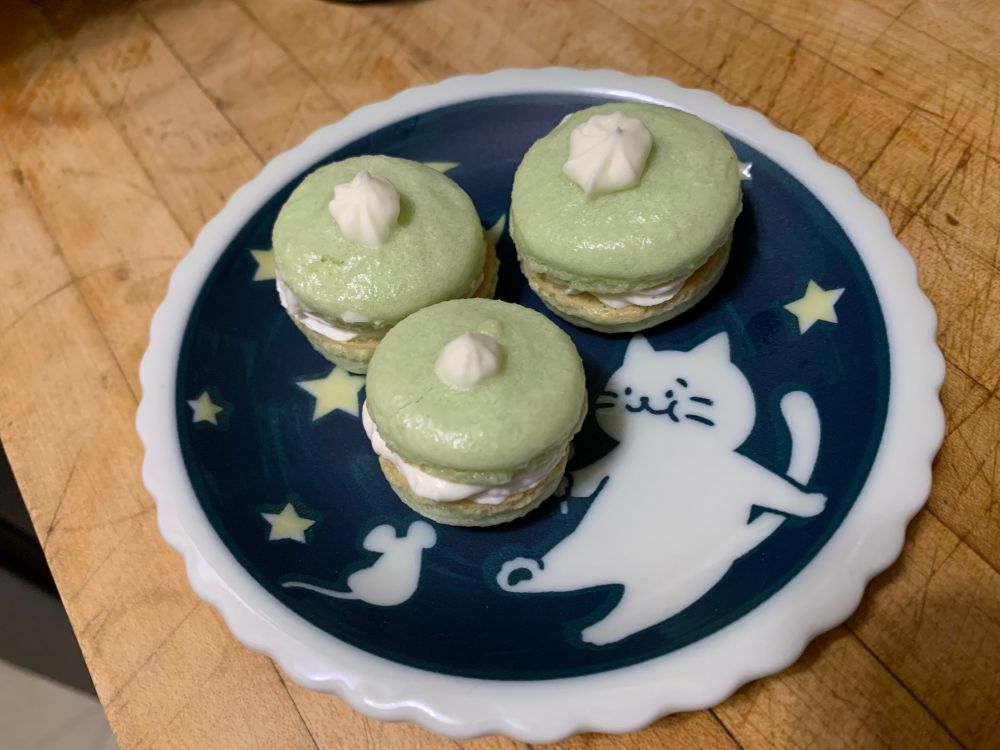Three green macarons on a small plate. The macarons are green with white buttercream filling and each have a dollop of buttercream on top. The plate has a cartoon cat and mouse, moon and stars on it.