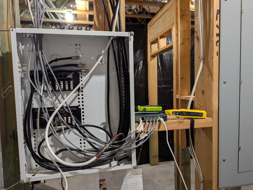 Home network setup in an unfinished basement. CAT 5 cables come out of an uncovered patchbox (unterminated) into a switch sitting beside it. A walkie talkie and diagnostic tool sit beside it.