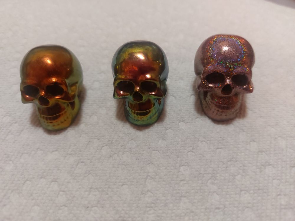 Three small resin skulls, maybe an inch high. The first one is a metallic red with green and gold highlights, the second is a very reflective pink/green/gold shifting color, and the third is pink holographic glitter. 