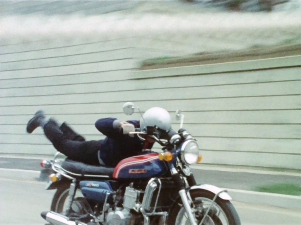 Kazami is on his bike and switched from sitting on it to lying down in a 'pass under the rope' maneuver 