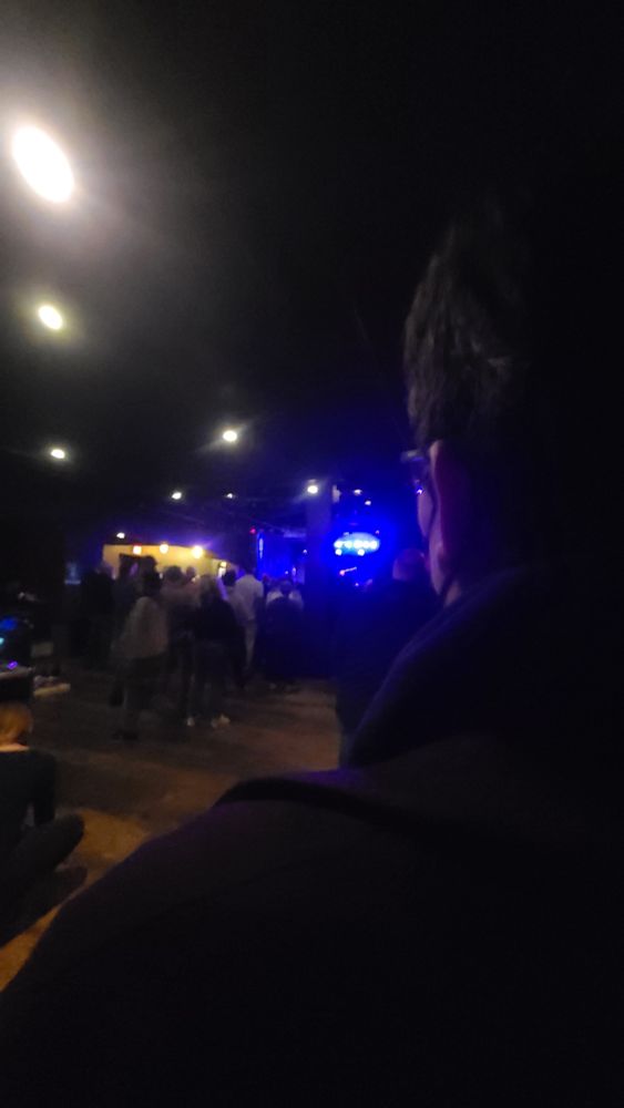 The back of a man's head, from behind his shoulder. You can see the arm of his glasses and the strap of his mask. He is looking at a crowd of people and a stage. A blue light above the stage illuminates a woman (Mary Bue) performing. 