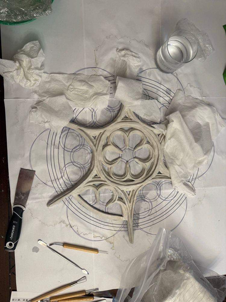 Image of a table where a white clay gothic rose window design is being built atop a printed design. Various clay tools are scattered around.