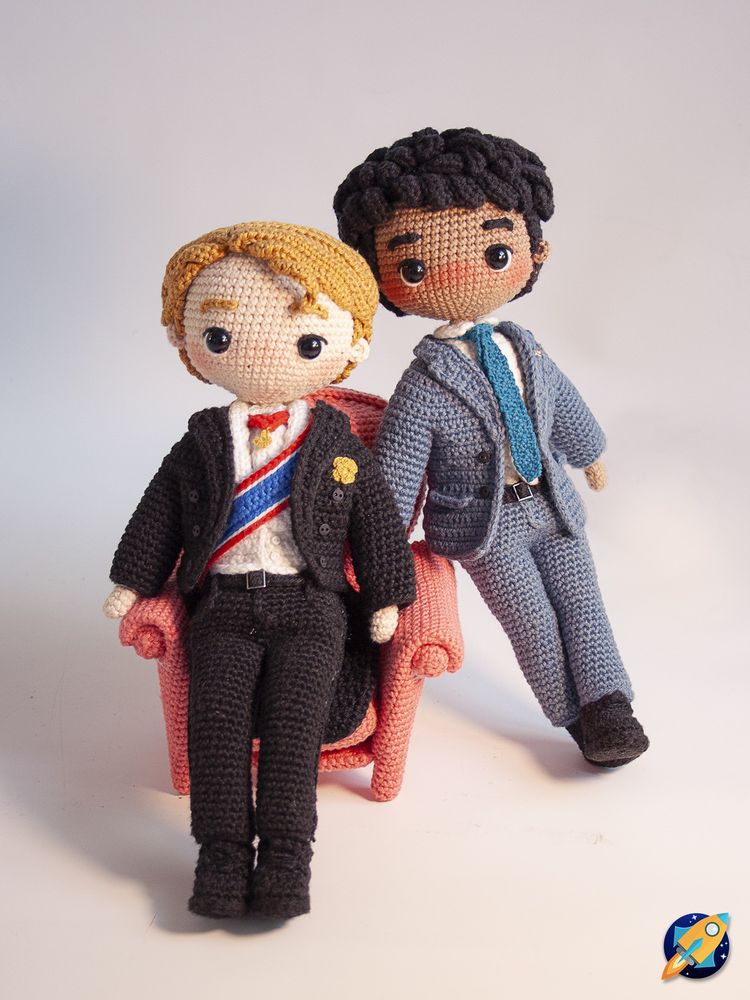Foto de dos amigurumis de Alex y Henry, los protagonistas de la película de "Rojo, Blanco y Sangre Azul". Henry lleva un frac negro con las galas propias de la realeza y está sentado en un sillón también hecho de crochet. Alex está a su izquierda, apoyado contra el sillón y con las piernas cruzadas y lleva un traje azul. Van vestidos como en los carteles promocionales de la película.