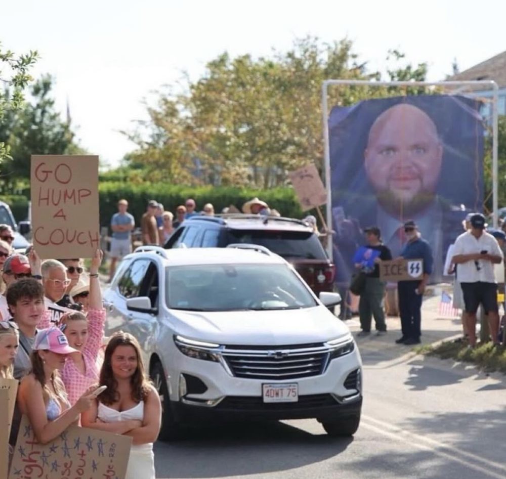 A sign protesting JD Vance reads “go hump a couch” and there is a larger than life poster of the bald JD Vance meme