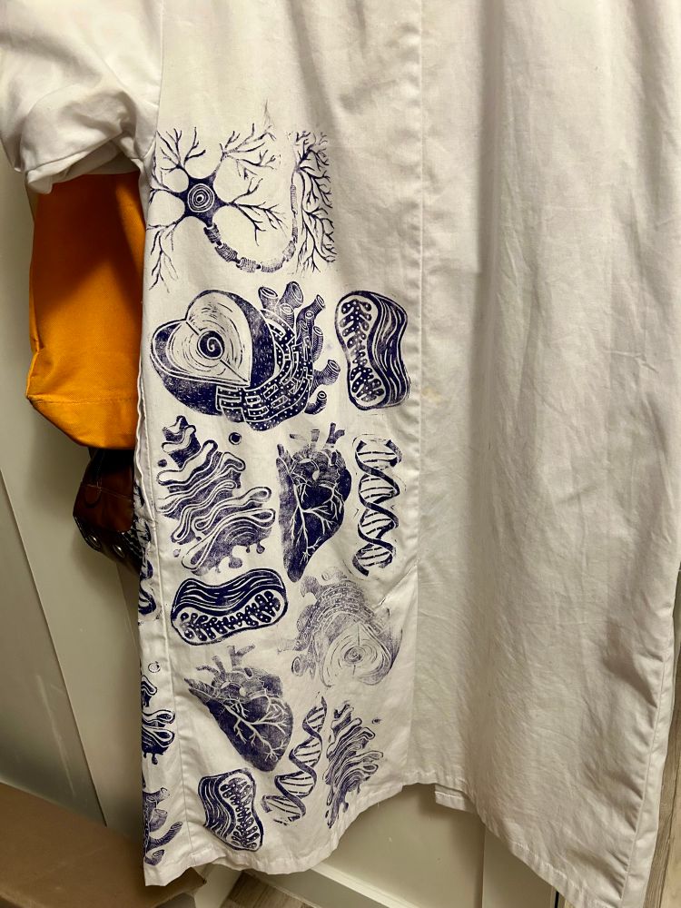 A lab coat partially covered in linocut block relief prints cut to look like organelles, DNA, neurons, and the heart. Printed in electric blue ink. 