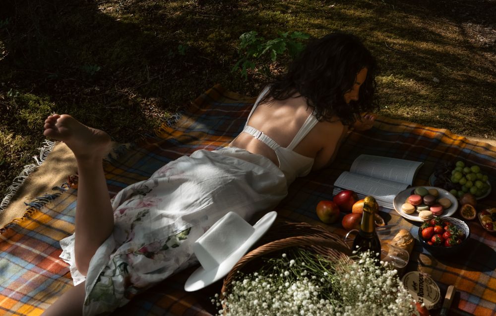 Danni is lying on her front, in dappled sunlight, on an orange and blue tartan picnic blanket. She is wearing a white mid-length floral dress and has bare feet, with one foot poised in the air. She is looking at an open book on the blanket beside her, her hair half-covering her face. The rest of the picnic blanket is filled with: a wicker basket holding white flowers, a white sunhat, champagne, French bread and cheese, fresh fruit, and a plate of macarons.