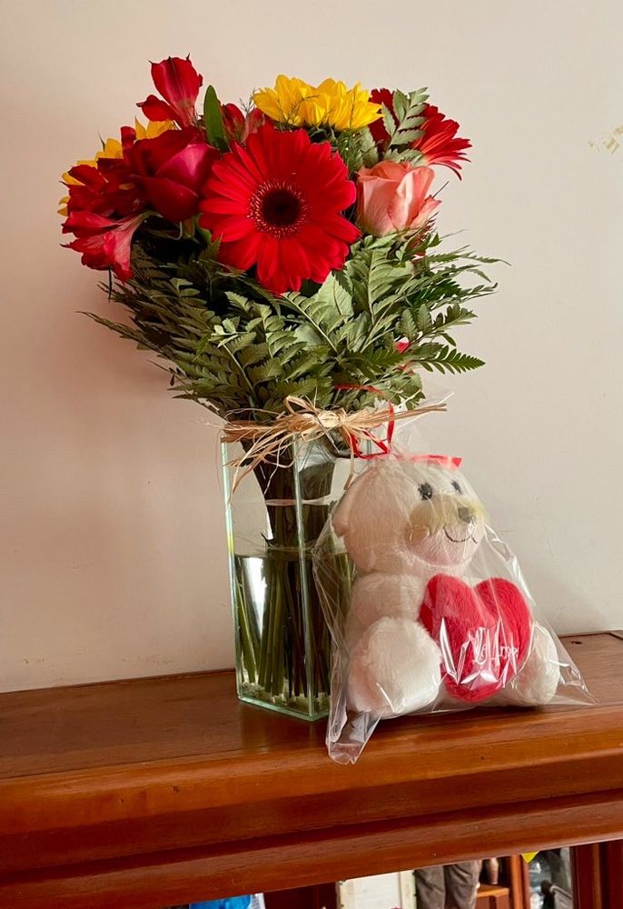Vaso de vidro com flores cor de rosa, vermelhas, amarelas e um ursinho de pelúcia. 