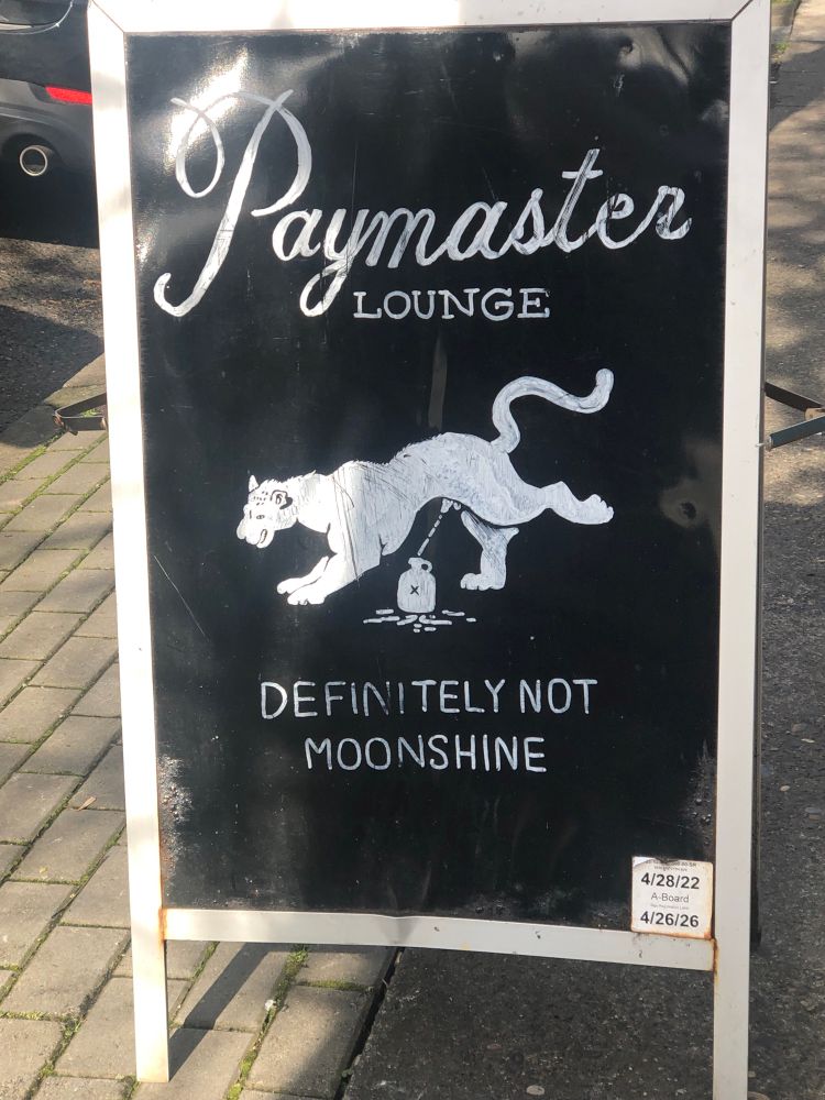 Sidewalk chalkboard sign for Paymaster Lounge featuring a white male cougar(?) or similar large cat kicking up its back leg and pissing into a moonshine jug, above the text “Definitely Not Moonshine” 