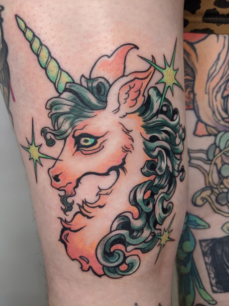 A back-of-the-thigh tattoo of a stylized unicorn portrait with tufted fur and a flowing sinuous mane. The unicorn’s body is rendered with soft peachy oranges while the mane has a cool, deep sea green palette. We used bright turquoise and chartreuse for the horn and the spiky 8 point stars accenting the piece. 