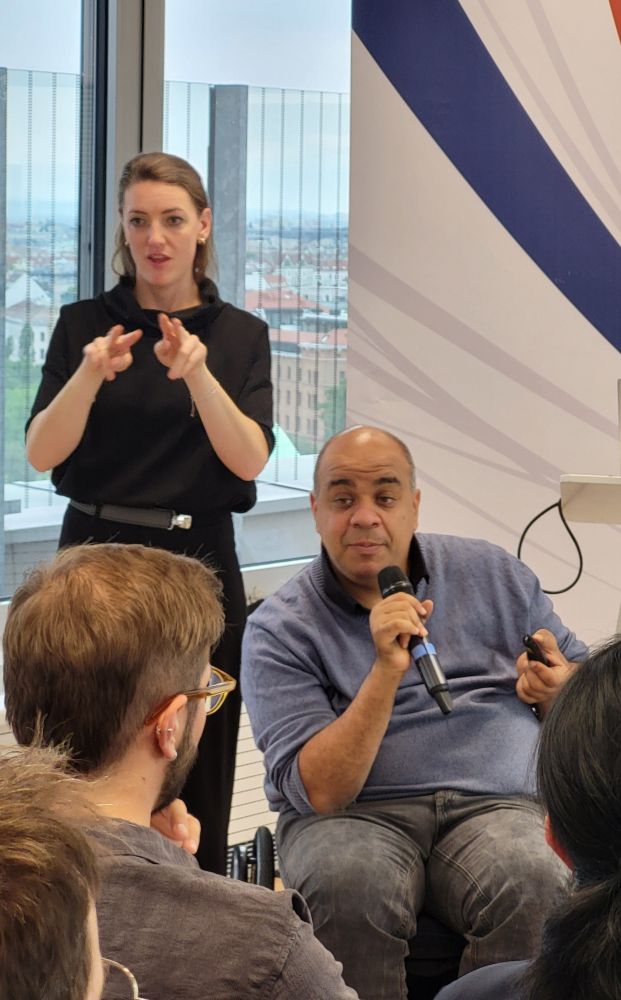 Shadi Abou-Zahra von Amazon hält seine Keynote mit einem Mikrofon in der Hand. Hinter ihm steht die Genärdendolmetscherin.