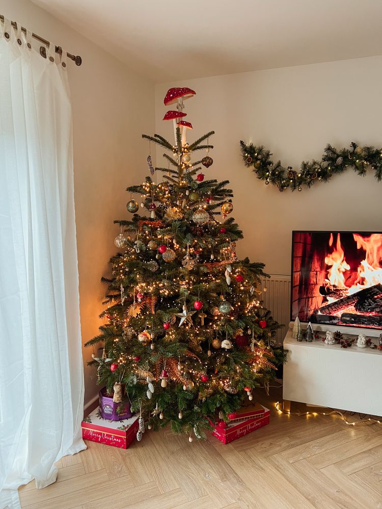A Christmas tree with red and gold ornaments and a large red mushroom on top. A tv screen to the side has a log fire video running. 