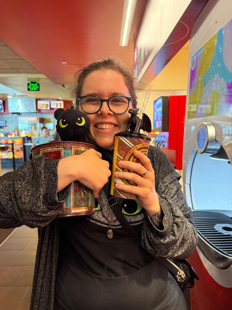 A picture of Robin Arnette about to see the live action How to Train Your Dragon movie. She has a toothless t-shirt, a HTTYD popcorn bucket with a toothless plushie inside, and a novelty HTTYD cup with a little toothless figurine on top. She has a HUGE smile, because of how much she loves both the movie and dragons in general