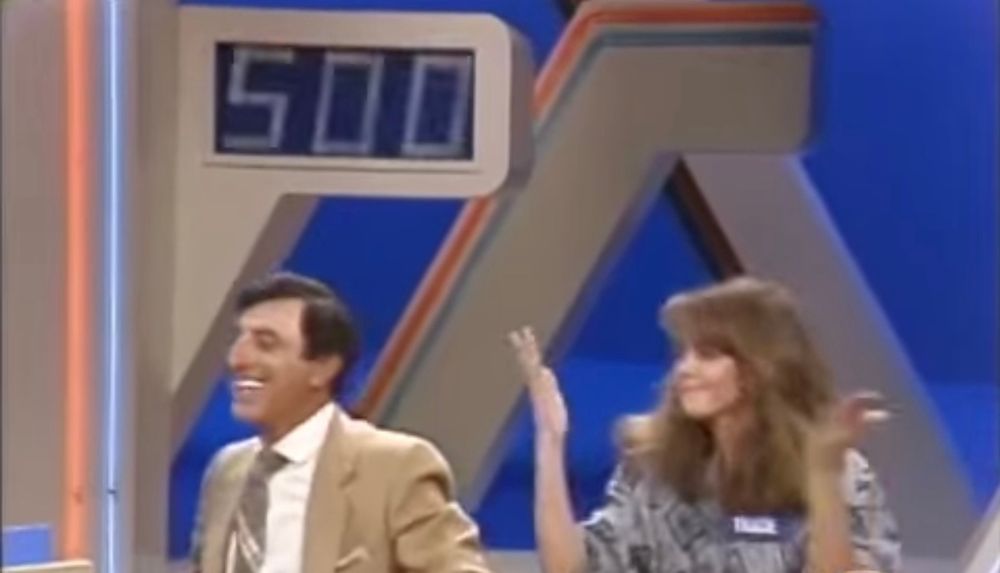 Jamie Farr smiles while a contestant who just won throws her hands up