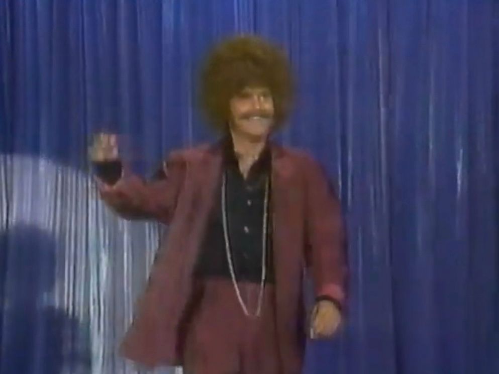 White man dressed in 70s disco garb and wearing an Afro wig 