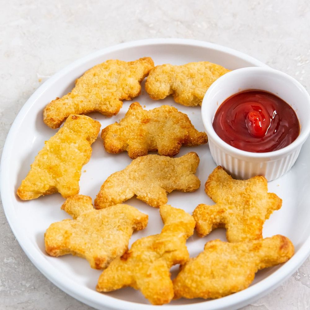Photo of a white plate on a gray surface. The plate contains many chicken nuggets shaped like dinosaurs and a bowl of ketchup. 