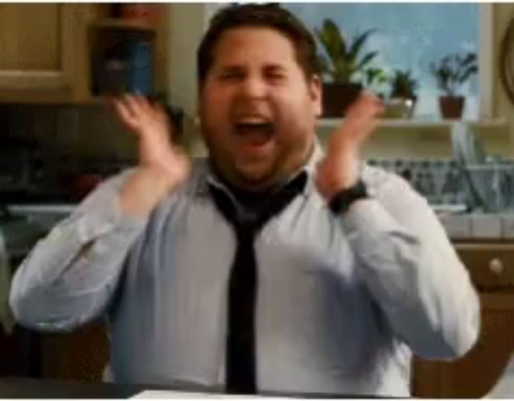 a man in a white shirt and tie is standing in a kitchen with his hands in the air .