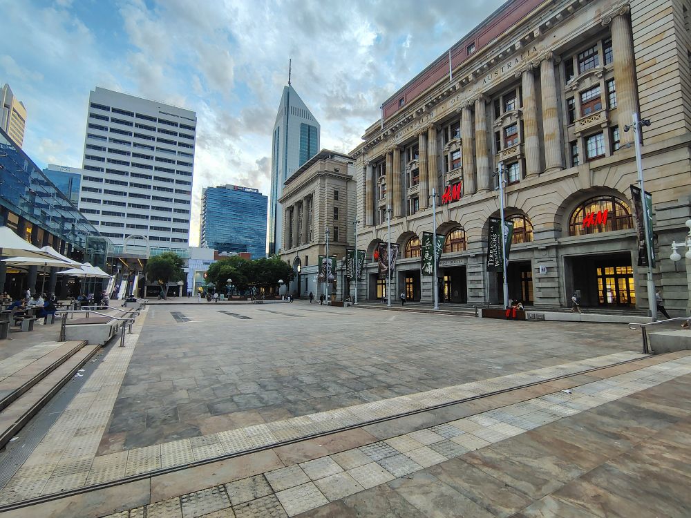 A paved plaza, with a large building to the side of it and taller buildings behind. There is seated dining under umbrellas along one edge but is otherwise bare and uninviting 