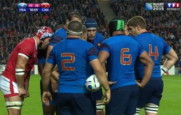 Photo du match entre la France et le Canada lors de la Coupe du Monde de Rugby 2015. On y voit un groupe d'une demi-douzaine de joueurs Français qui mettent en place leur tactique pour l'action de jeu à venir. Juste à côté d'eux et penché vers le groupe comme s'il participait, on voit Jamie Cudmore, joueur du Canada, qui s'incruste pour écouter la conversation.