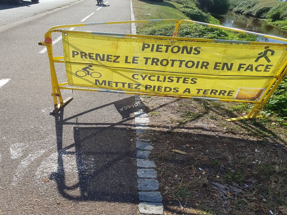 Photo d'une piste cyclable en partie barrée par un assemblage de trois barrières soutenant un tissu jaune sur lequel sont fixées deux bandeaux de travaux imprimés avec le texte "Piétons prenez le trottoir en face" et "Cyclistes mettez pieds à terre".