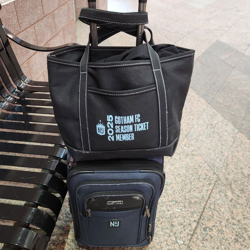 A 2025 GothamFC black tote bag sits on top of a dark blue carry on suitcase.