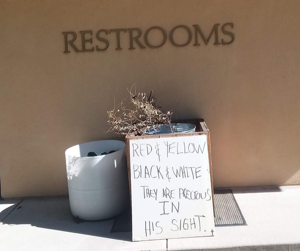A large handwritten sign leans against a rubbish can outside a cemetery public restroom. The sign reads: 