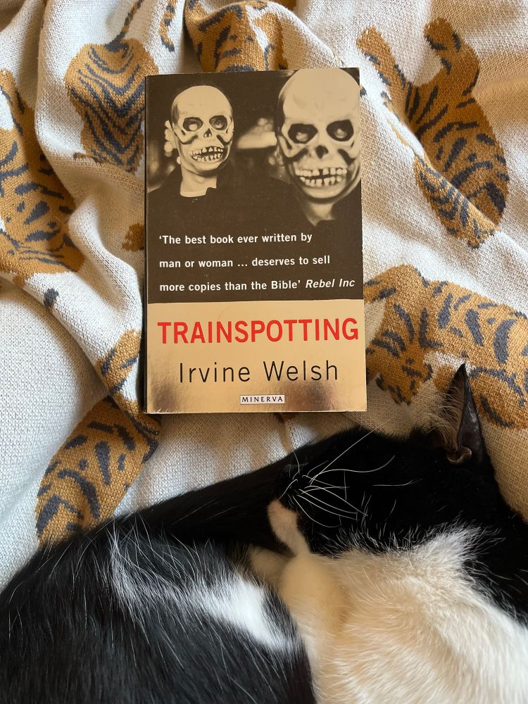 Paperback copy of Trainspotting next to a tuxy cat; blurb says “the best book ever written by man or woman…deserves to sell more copies than the Bible”