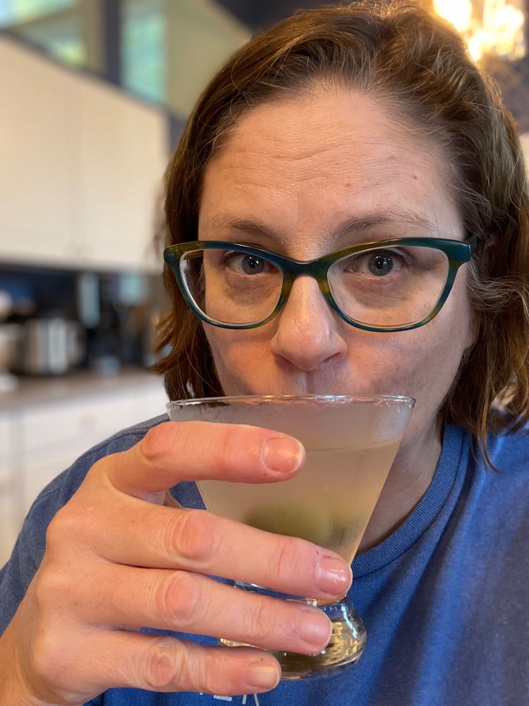 An exhausted middle-aged white woman in green glasses, gratefully sucking down an ice cold dirty martini. 