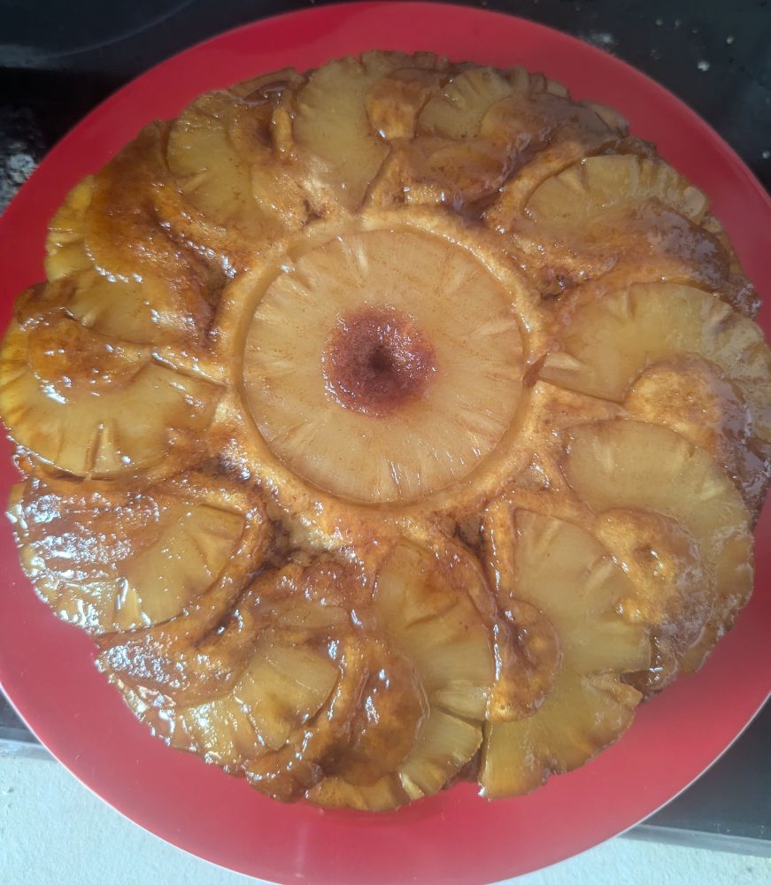 The image shows a beautifully baked pineapple upside-down cake. The cake has a rich caramelized glaze with pineapple rings arranged in a circular pattern, with one pineapple ring in the center and smaller slices radiating outward. The golden-brown cake beneath complements the shiny, juicy pineapple topping. Alas, the cake showing through the inner pineapple ring looks like an anus.