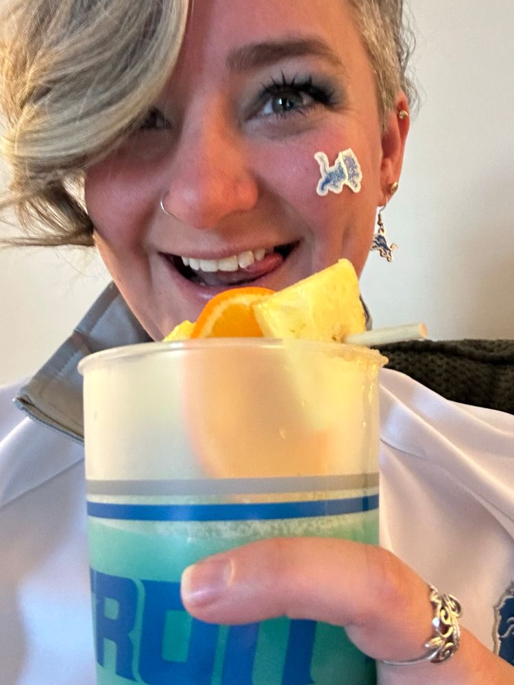 Selfie of me smiling with my tongue slightly out to the side. I’m wearing a lions logo sticker on my cheek, lions logo earrings and a lions zip up. I’m holding a lions cup with Blue Drink 2.0 in it. I have a silver hoop nose ring and a swirly leaf ring on my index finger. 