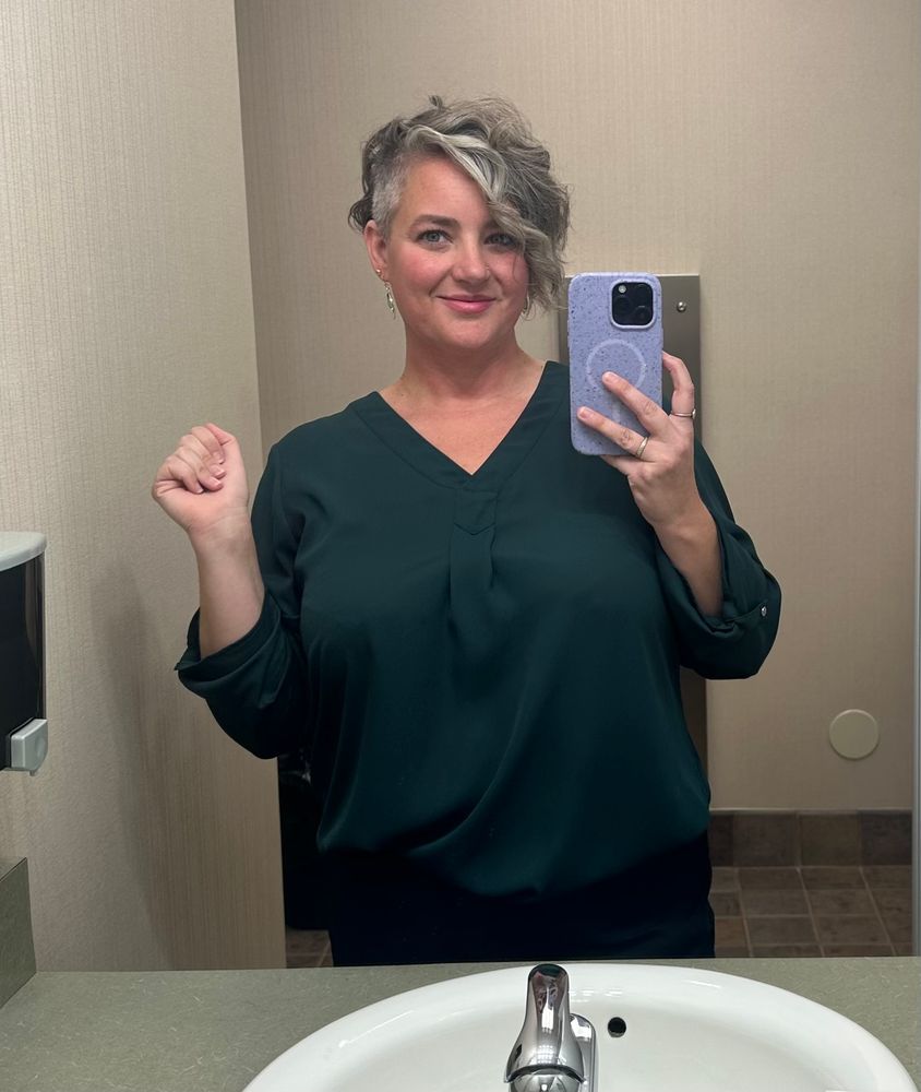 Selfie of me in my office bathroom mirror. I’m wearing a flowy dark emerald/ jungle green top and am holding my phone up wit the millennial hand pose of my pinky underneath and fingers supporting. I’m smiling and have my other hand raised in a lovely closed fist because I don’t know why. You can see part of the sink in the bottom of the frame and part of the soap dispenser on the left. 