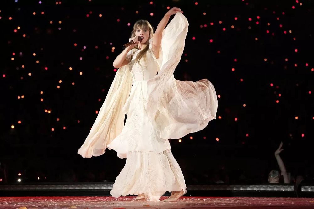 Picture of Taylor Swift on stage in the cream twirly gown at the eras tour. The background is dark and you can see glowing bracelets behind her 