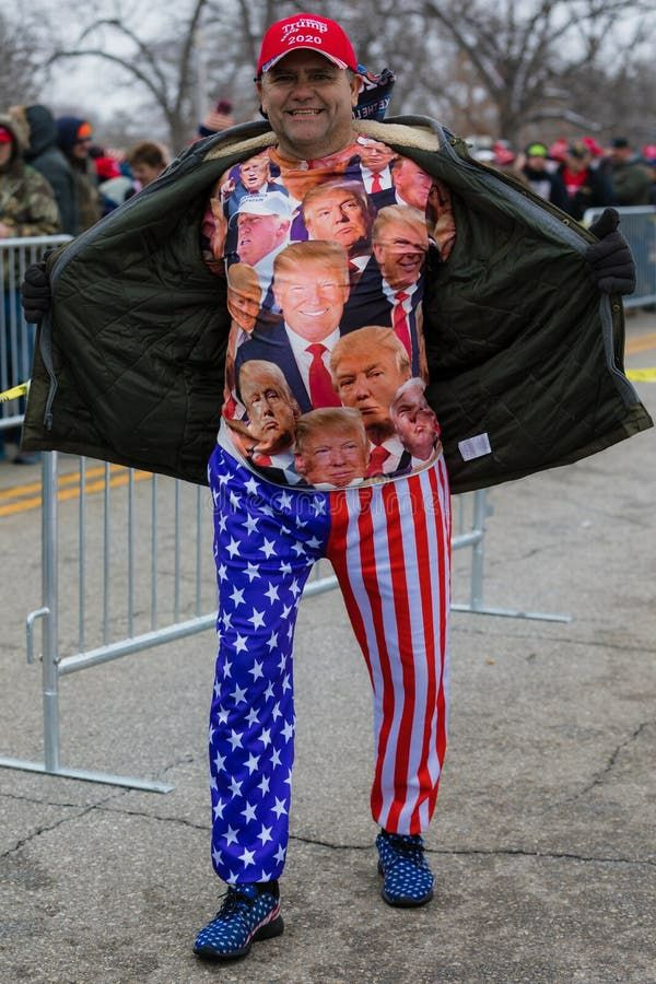 The picture shows a grinning man opening his jacket to reveal a shirt printed with a collage of portraits of Donald Trump. He wears a MAGA cap and stars-and-stripes trousers. He stands in front of a large crowd behind a fence.