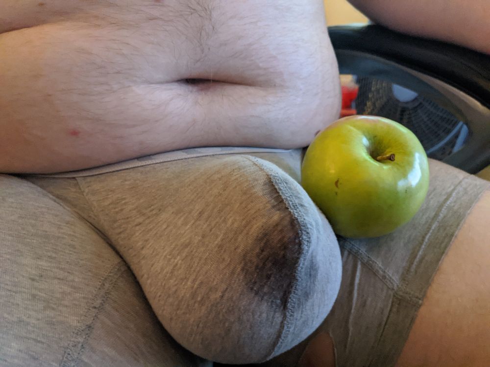 A man sits in bulging underwear wet with precum. A granny Smith Apple rests against the bulge  which is clearly larger than the apple. 