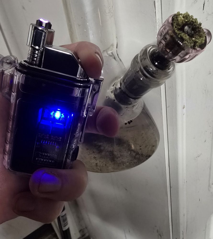 A hand holds a funky electric arc lighter designed to look like an electronic zippo with its shiny metal housing in a plastic case. The case is a part of the housing. It has a flip top similar to a zippo to cover and seal the electronic components inside. 

On the side facing the viewer, a blue LED shines through a window on the side, indicating activity. The circuit board and chips of the electronics show through the same window cut in the metal, protected by the clear plastic case. 

At the top, a button replaces the usual striker and fuel lever, activating the two electric arcs at the end of a retractable gooseneck conduit, placed where a flame would usually ignite.

This overall lends the lighter a somewhat "cyberpunk" aesthetic. 

Behind that, the bottom of a hanging bong can be seen, its green contents partly burned. Presumably by the electronic lighter being shown off in the image. 