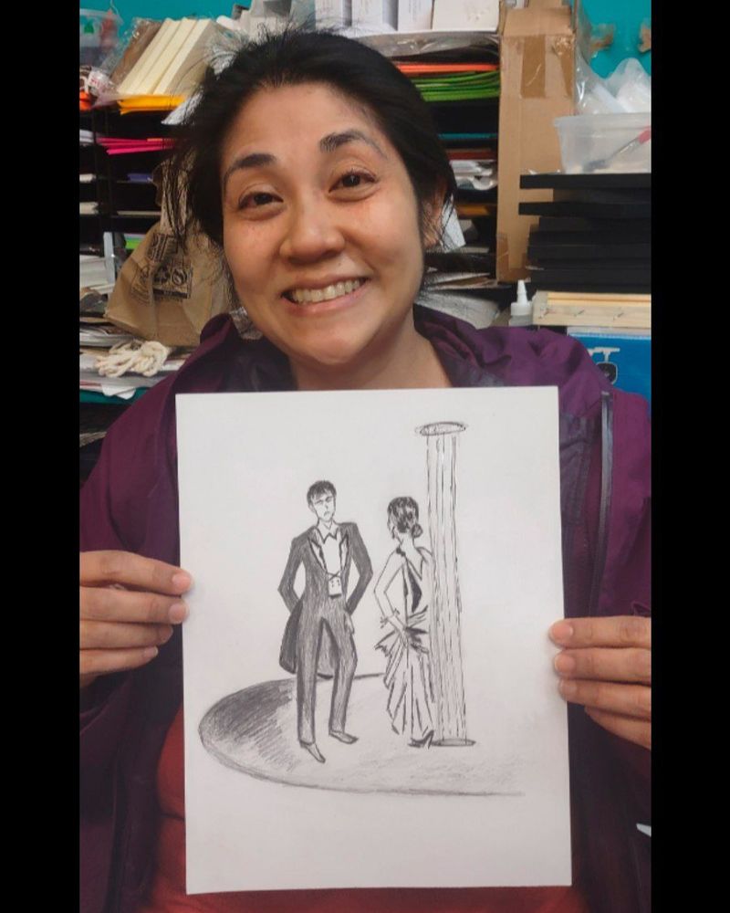 Photo of a smiling person holding up a pencil drawing of two characters in a ballroom setting.