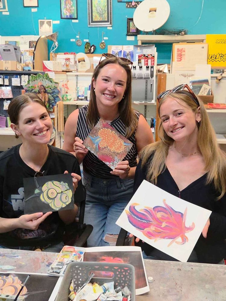 Three people smiling at camera, each holding their own small painting of a colorful sea slug.